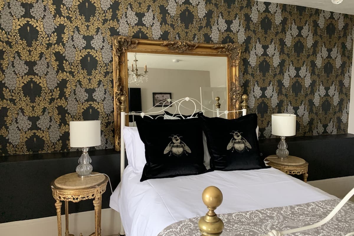 Une chambre à coucher avec un grand miroir décoratif, un lit recouvert de draps blancs, deux tables de chevet avec des lampes et un papier peint à motifs floraux.