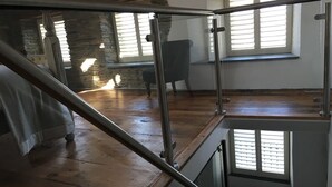 Una habitación con piso de madera, una escalera con barandillas de vidrio, una pared de piedra y una ventana con persianas.