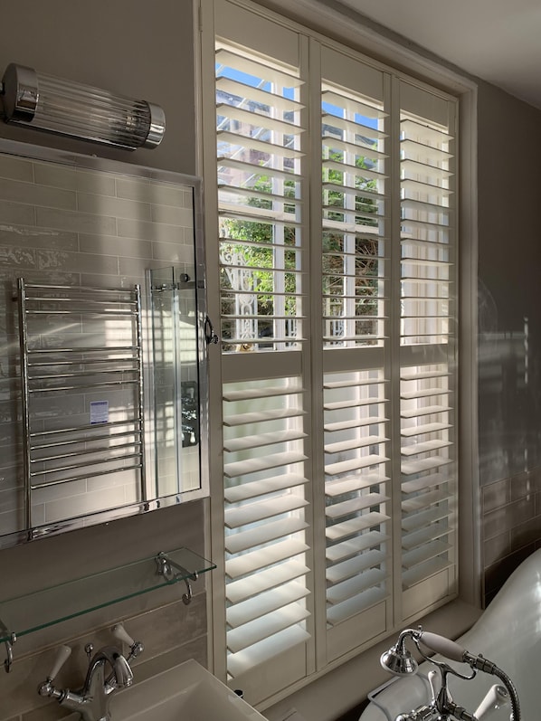 A bathroom with a glass shower enclosure, a white sink, and a large window with blinds.