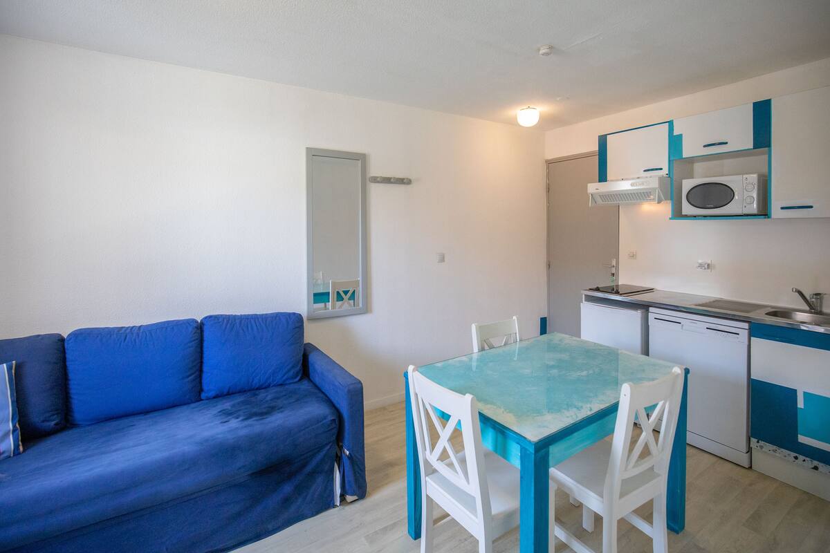 A compact living space with a blue sofa, a glass-top dining table, white chairs, a microwave, a small sink, and a compact kitchenette.