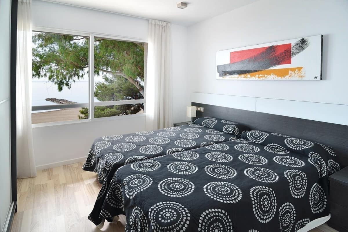 A bedroom with a large window, a bed with patterned bedding, a dark headboard, and a piece of abstract art on the wall.
