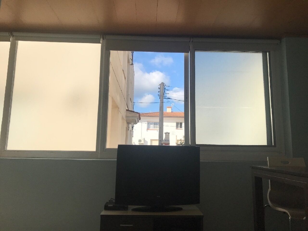A room with a television on a stand, a window with a view of buildings, and a cabinet to the right.