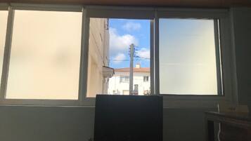A room with a television on a stand, a window with a view of buildings, and a cabinet to the right.
