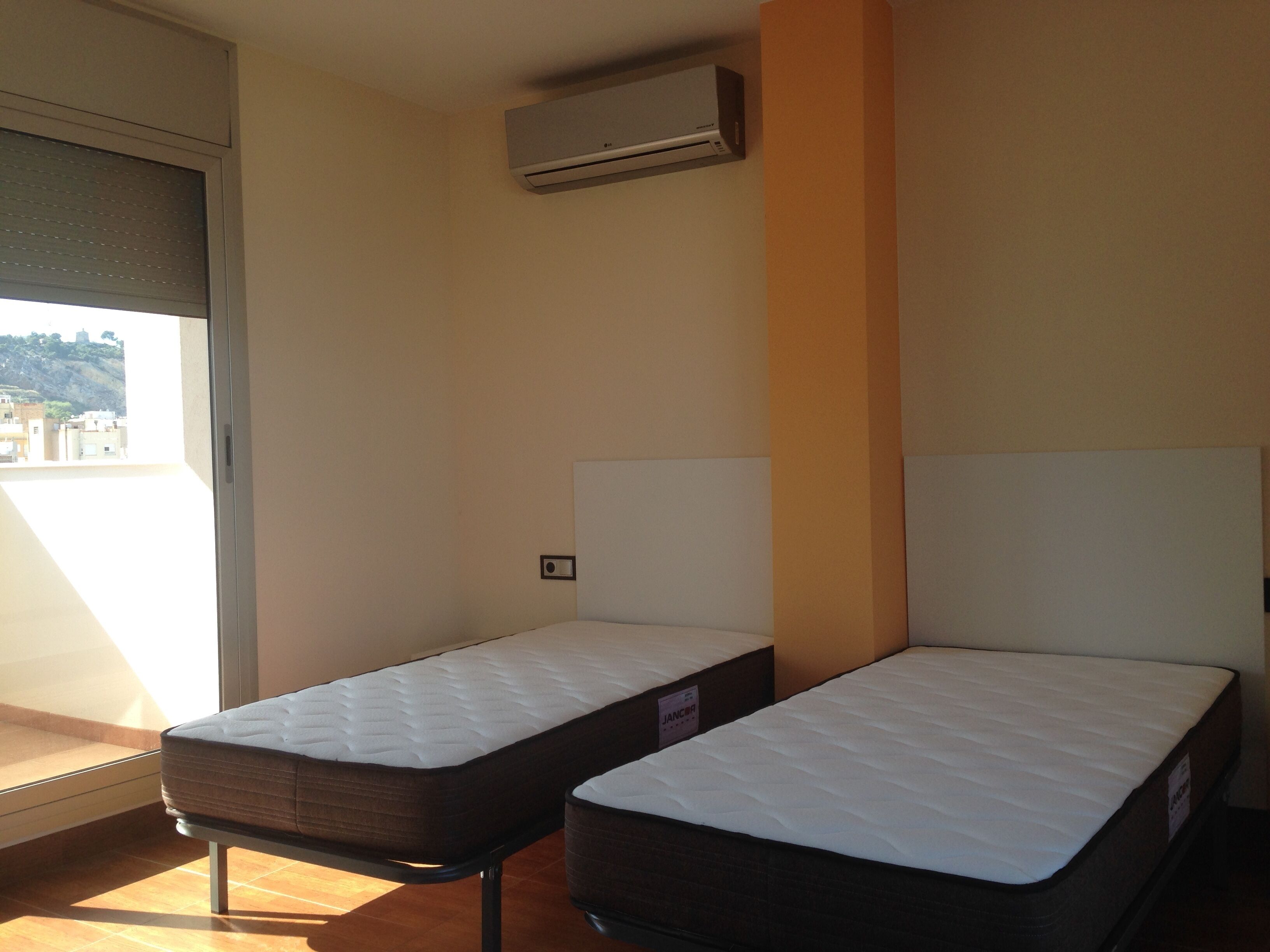 Two single beds with headboards, a window with blinds, and an air conditioning unit on the wall.