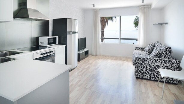 A modern kitchen with a microwave, stove, and TV, a black and white patterned sofa, and a large window with a view of the sea. - Apartamentos Ornis (Sant Carles de la Rapita)