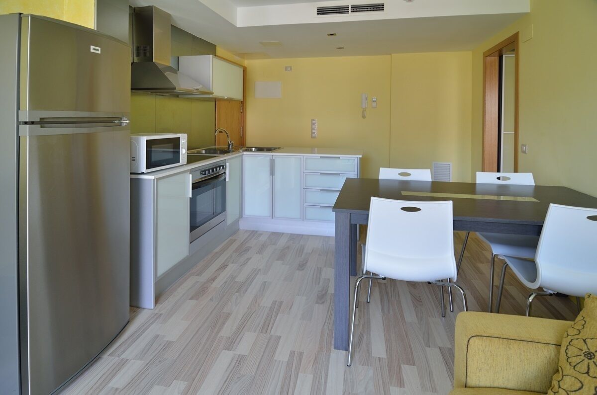 Cocina compacta con refrigerador, microondas, estufa y gabinetes blancos. Hay una mesa de comedor con sillas y un sofá amarillo.