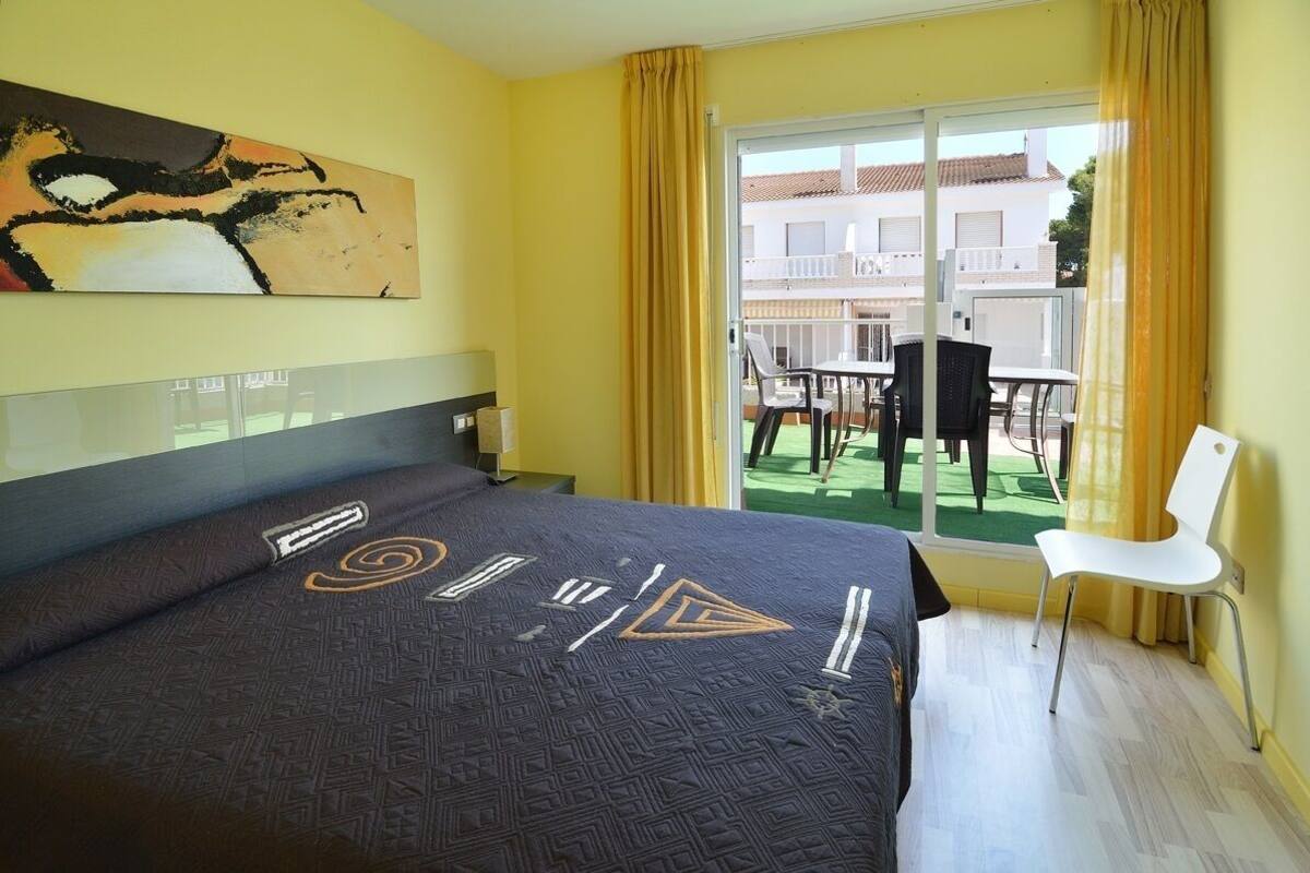 A hotel room with a bed, a painting on the wall, a balcony with chairs, and a view of buildings outside.