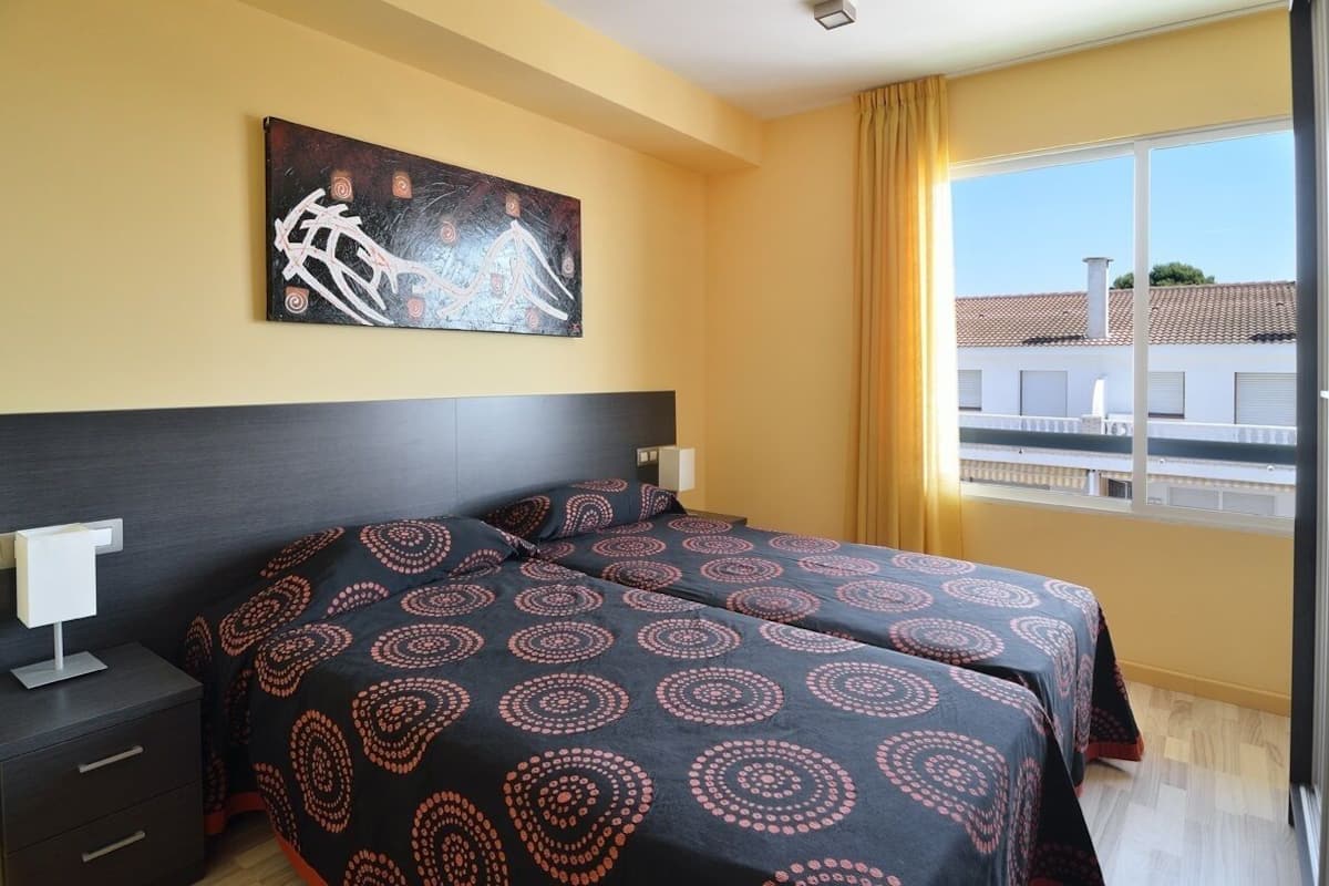 A hotel room with a bed, a nightstand, a lamp, a painting on the wall, and a window with a view of buildings.