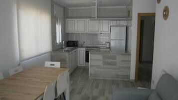 Una cocina compacta con armarios blancos, un microondas y un refrigerador. Hay un comedor con una mesa y sillas de madera.