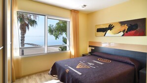 A hotel room with a bed, a nightstand, a lamp, a painting, and a window with a view of the sea and palm trees. - Apartamentos Ornis (Sant Carles de la Rapita)