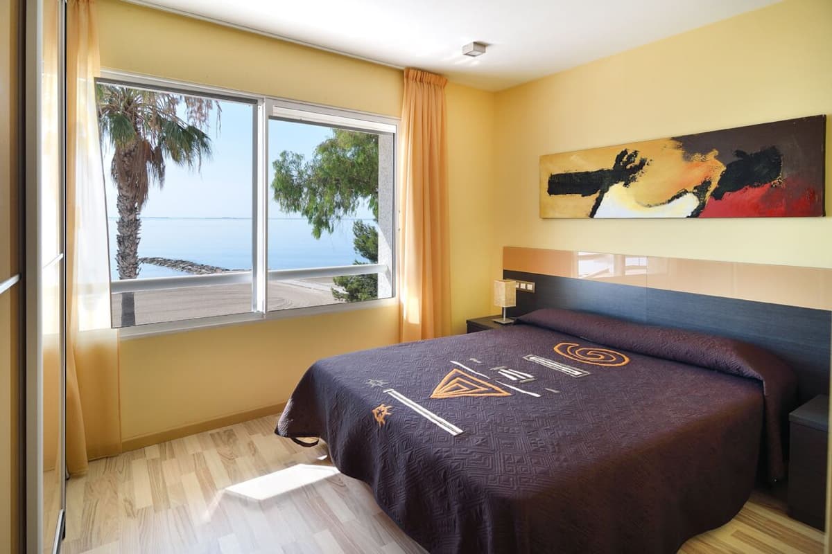 A hotel room with a bed, a nightstand, a lamp, a painting, and a window with a view of the sea and palm trees.