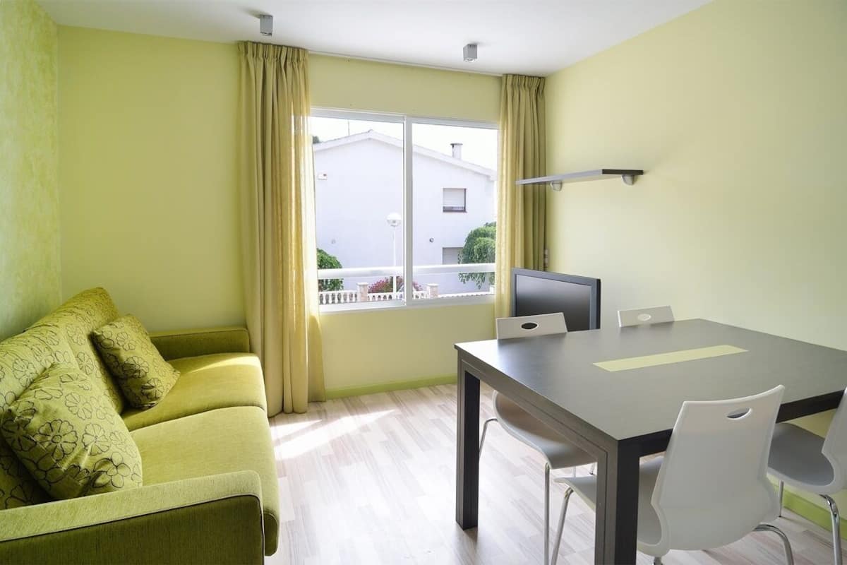 A room with a green sofa, a dining table with chairs, a TV, and a window with curtains.