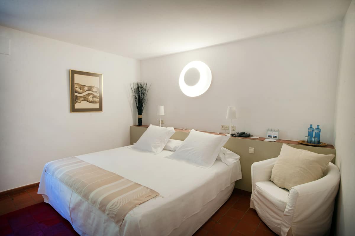 Un lit bien fait, avec du linge de lit blanc, une lampe de chevet et une petite table sur laquelle sont posés des articles de toilette.