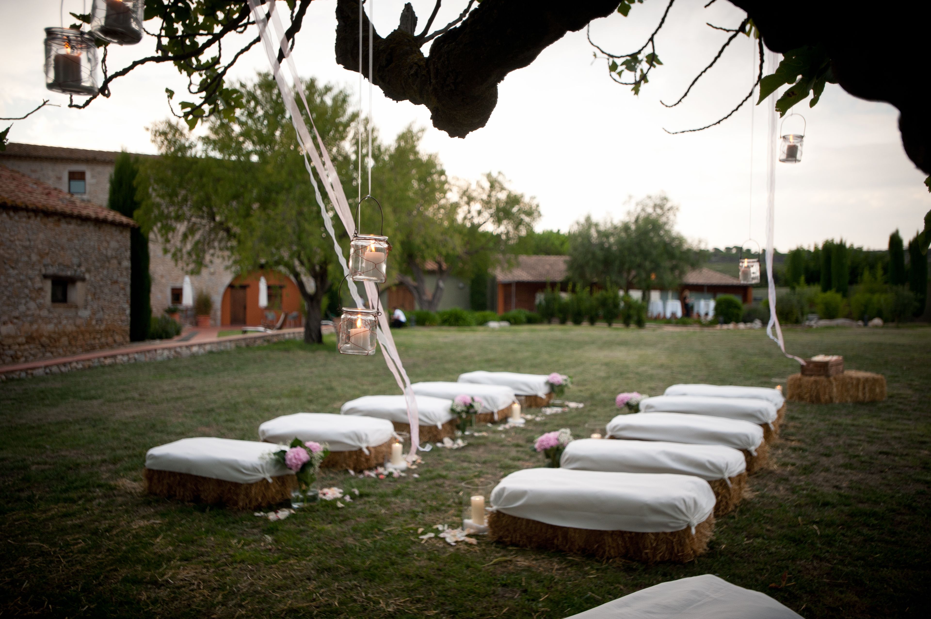 A rustic outdoor wedding setup with hay bales arranged in rows, each with a white cushion and a candle.