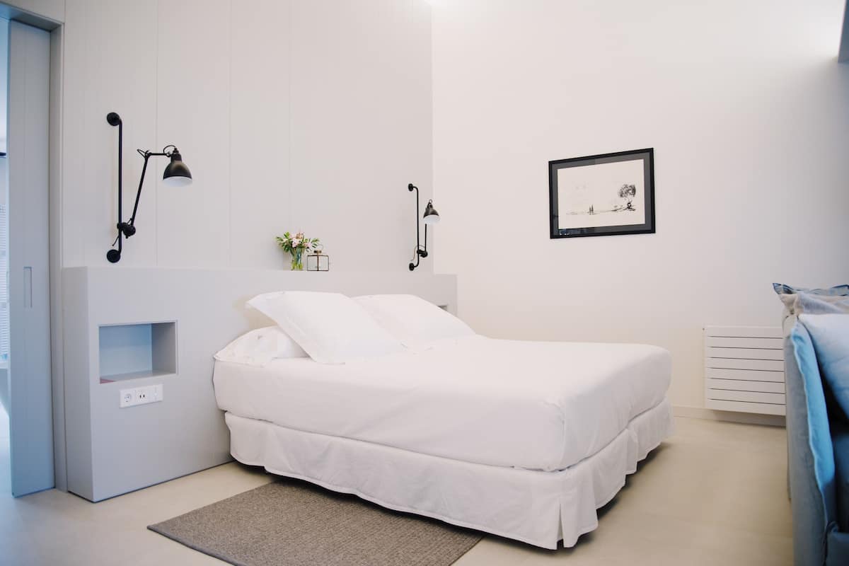 Une chambre moderne avec un lit blanc, une table de chevet, une lampe fixée au mur et un tableau encadré accroché au mur.