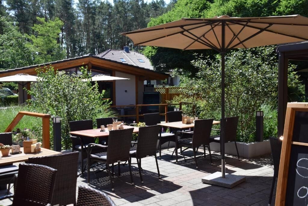An outdoor patio area with tables and chairs, surrounded by greenery and a building in the background.
