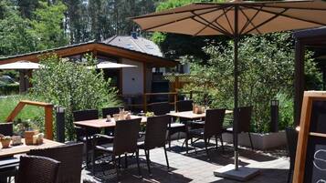 An outdoor patio area with tables and chairs, surrounded by greenery and a building in the background.