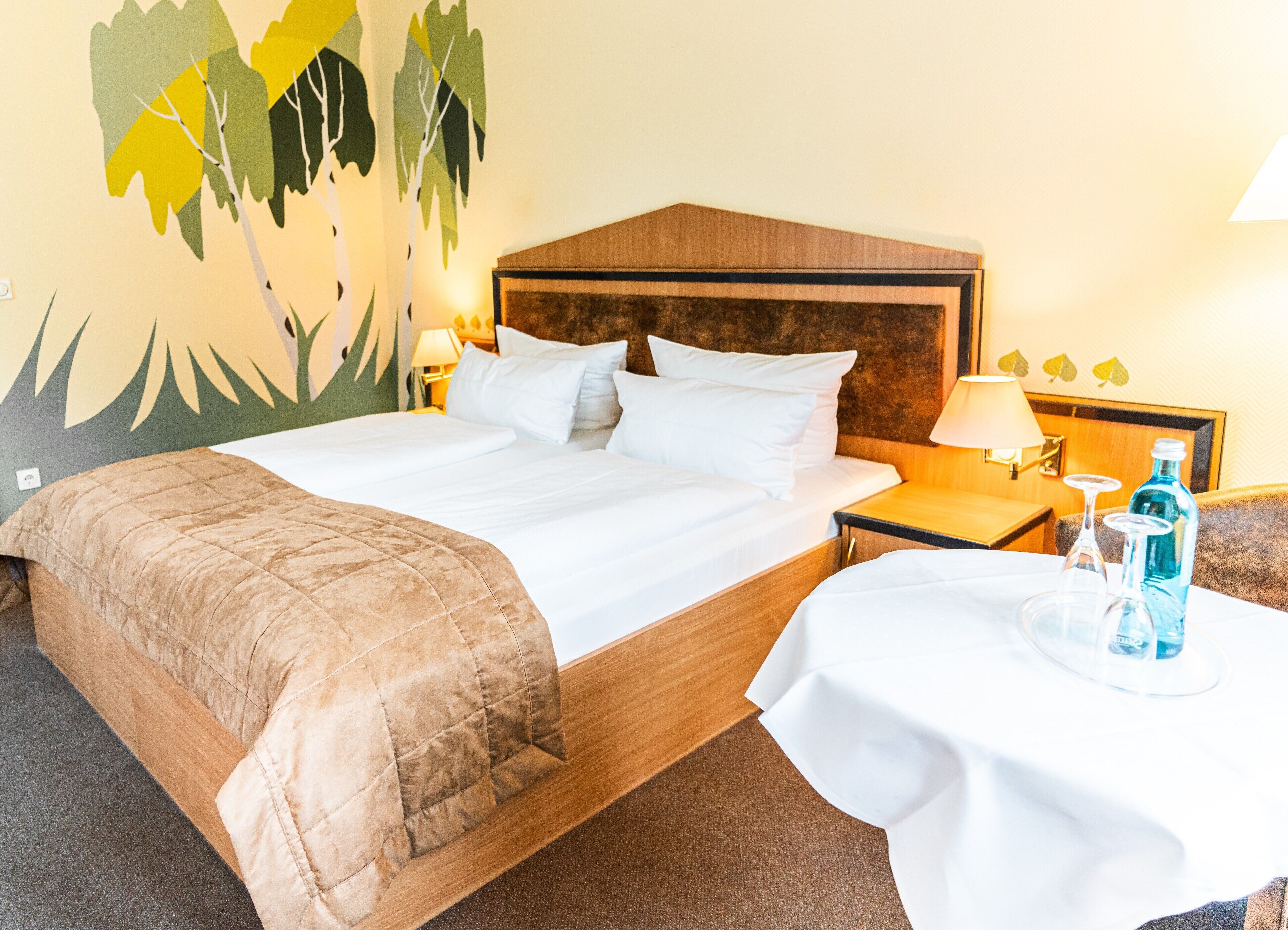 A hotel room with a large bed, a wooden headboard, a bedside table with a lamp, and a small table with a bottle and glasses.