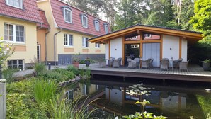 A building with a red roof, a wooden deck with outdoor seating, and a pond with lily pads. - Hotel Zum Leineweber (Burg (Spreewald))
