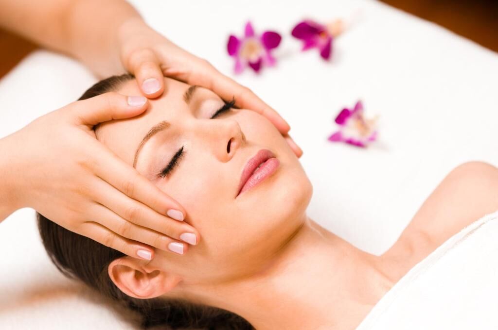 Eine Person liegt mit den Händen auf dem Kopf, möglicherweise erhält sie eine Massage, im Hintergrund sind violette Blumen zu sehen.