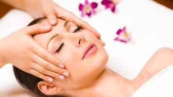Eine Person liegt mit den Händen auf dem Kopf, möglicherweise erhält sie eine Massage, im Hintergrund sind violette Blumen zu sehen.