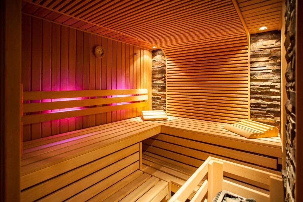 A sauna with wooden benches and slatted walls, a clock, and a stone wall.