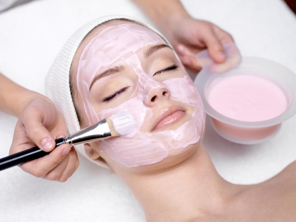 A person receiving a facial treatment with a brush and a bowl of pink substance.