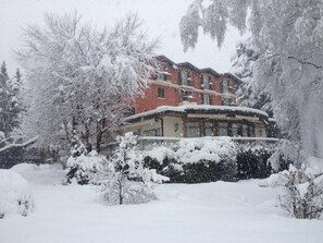 A building with a snow-covered roof and surrounding trees, indicating a winter setting. - Albergo Milano (Alta Valle Intelvi)
