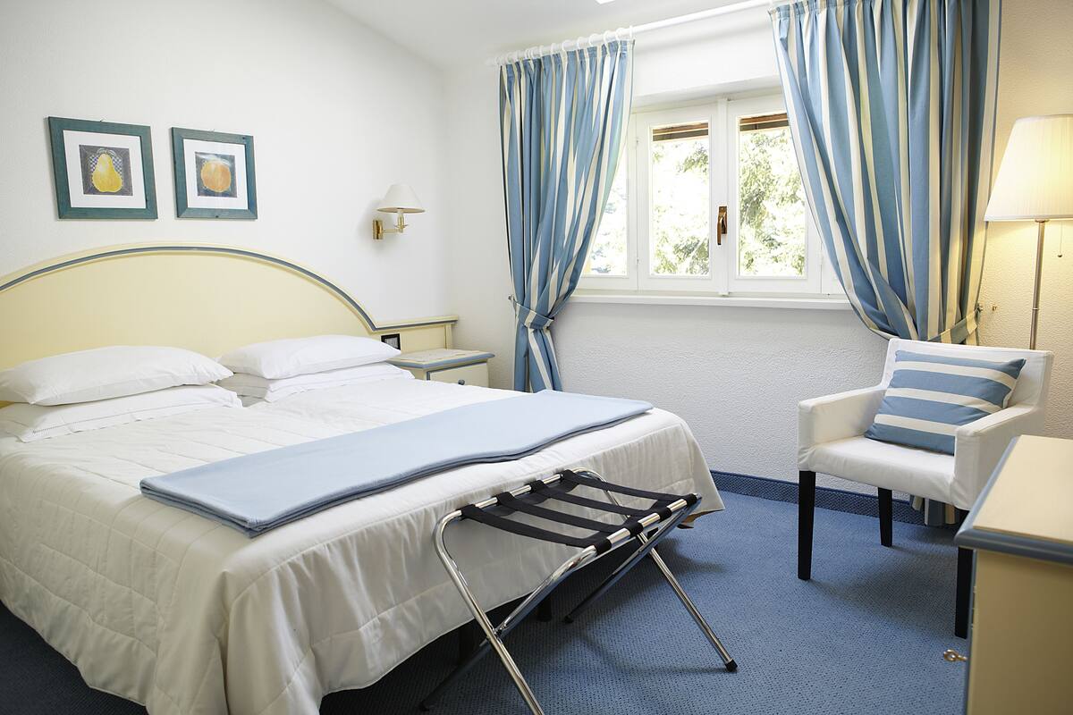 A neatly made bed with a blue blanket, a chair with a striped pillow, and a bedside table with a lamp.