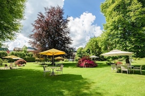An outdoor patio area with tables and chairs under umbrellas, surrounded by lush greenery and trees. - Albergo Milano (Alta Valle Intelvi)