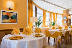 A dining area with round tables set for a meal, featuring yellow napkins and gold cutlery. - Albergo Milano (Alta Valle Intelvi)