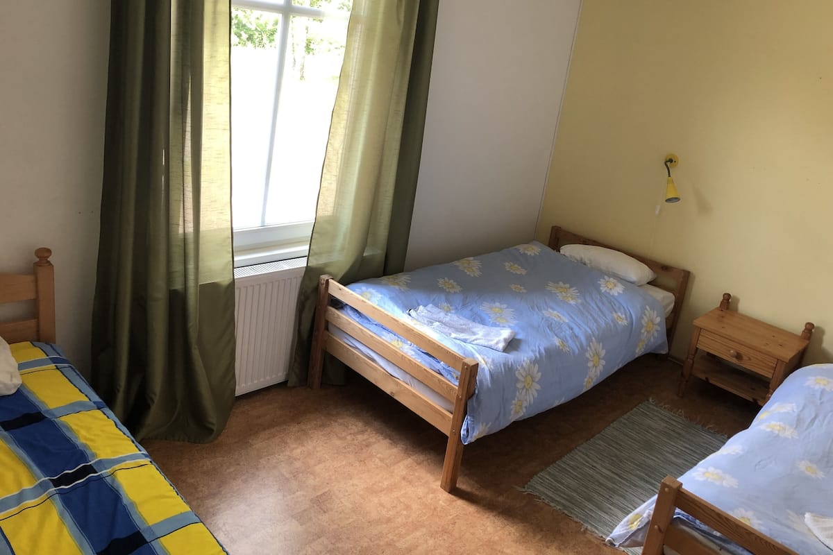 A bedroom with a single bed, a wooden nightstand, a radiator, and a window with curtains.