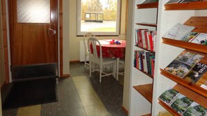 Una habitación con puerta de madera, ventana con vistas a árboles, una mesa con mantel rojo y una estantería llena de libros.