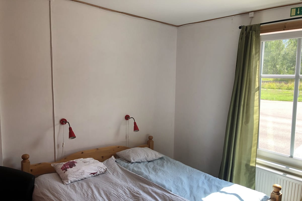 A single bed with a wooden headboard, two pillows, a red wall lamp, and a window with a view of greenery.
