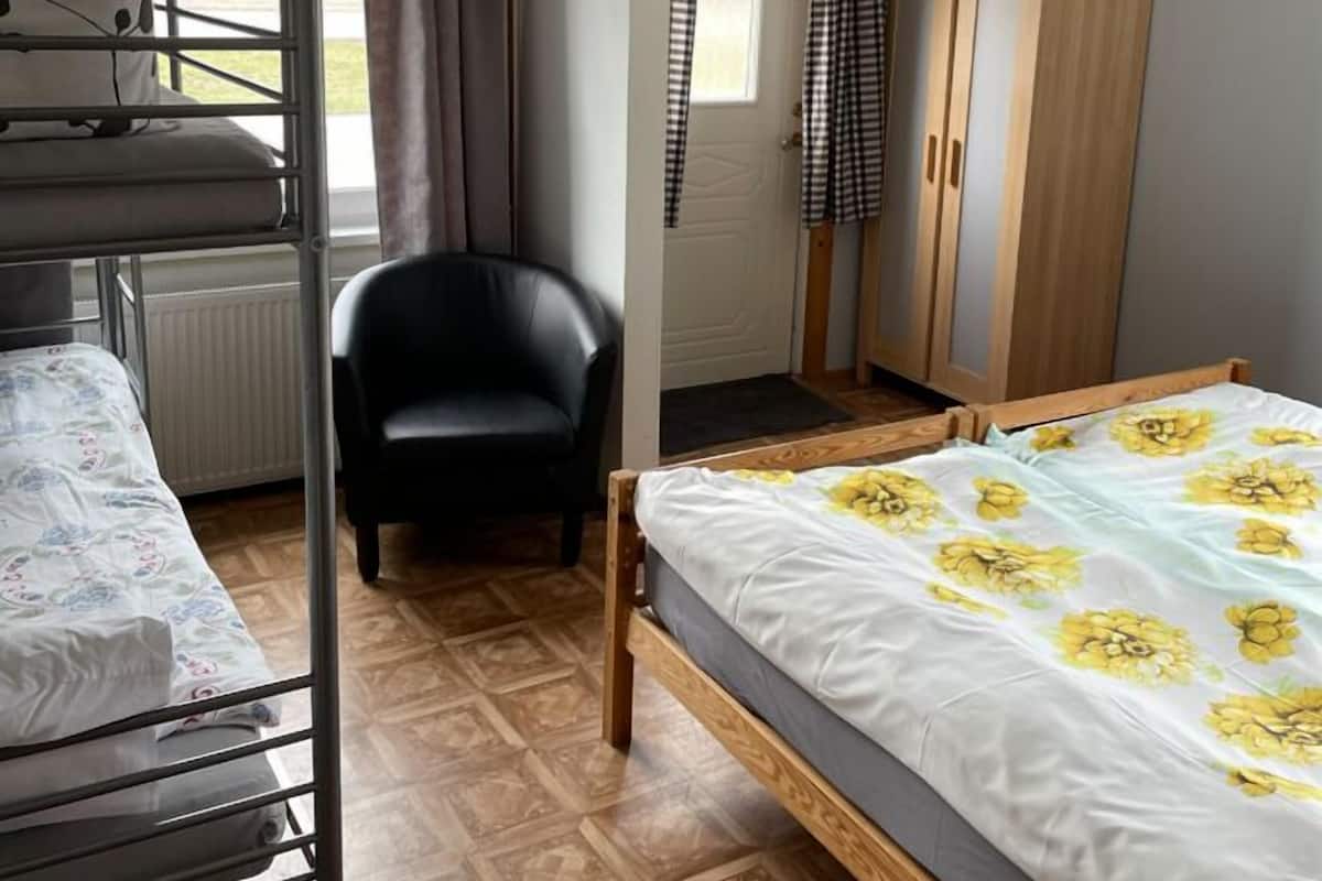 A dormitory room with bunk beds, a single bed, a chair, a wardrobe, and a window with curtains.