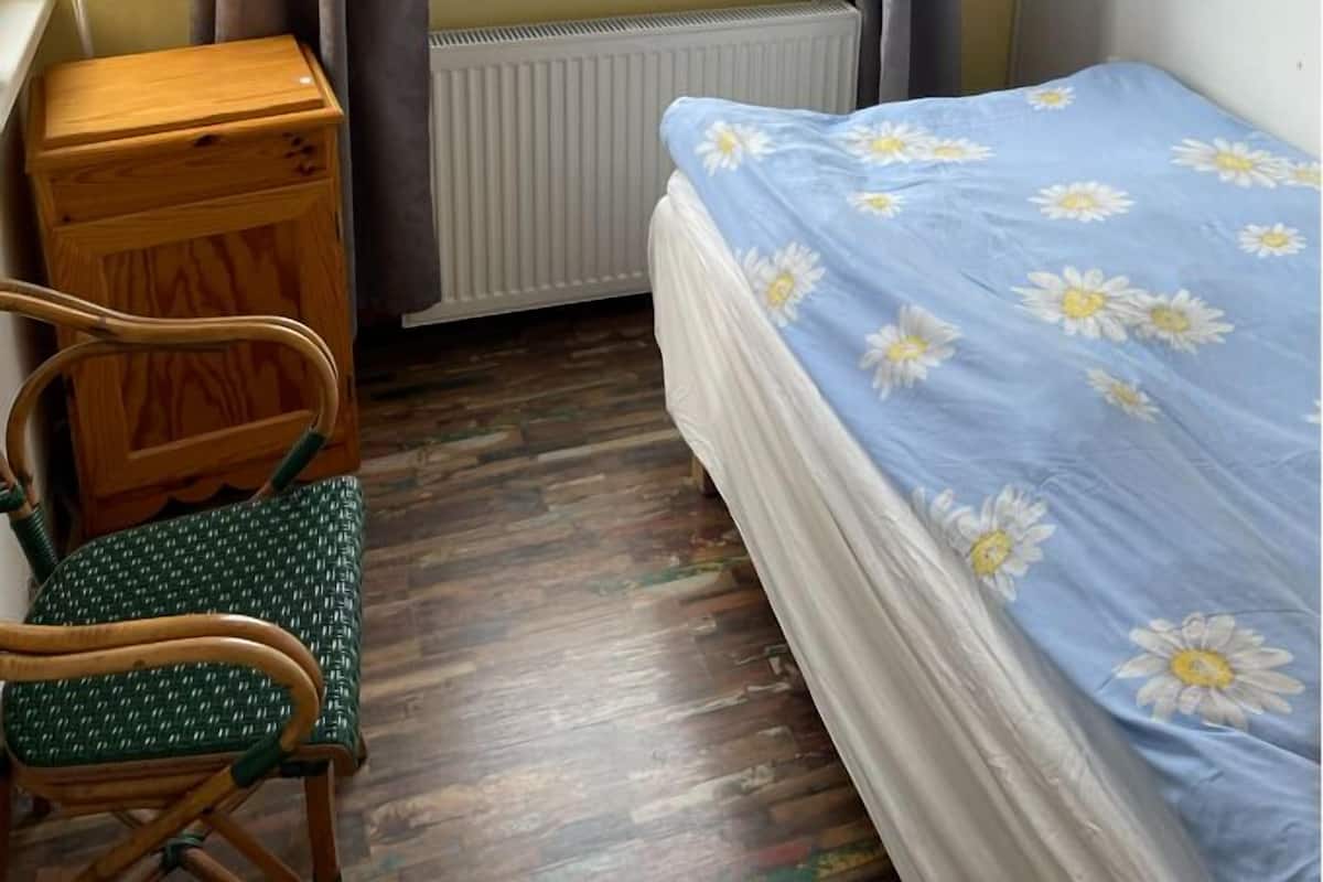 A room with a bed, a chair, a desk, a radiator, and a window with curtains.