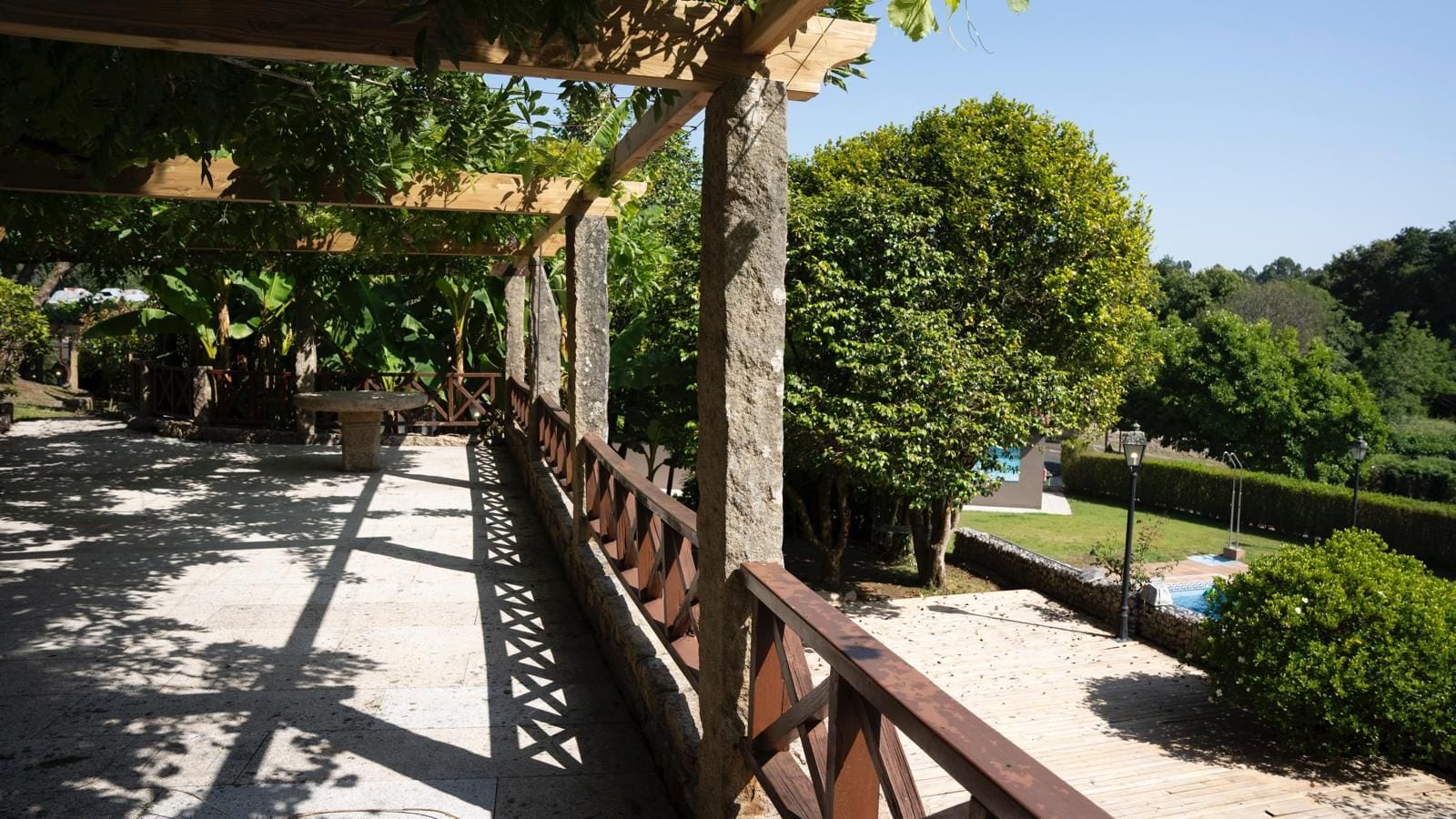 Zona exterior coberta amb barana de fusta, pilars de pedra i vistes a un jardí paisatjistic amb una piscina.