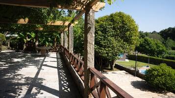 Zona exterior coberta amb barana de fusta, pilars de pedra i vistes a un jardí paisatjistic amb una piscina.
