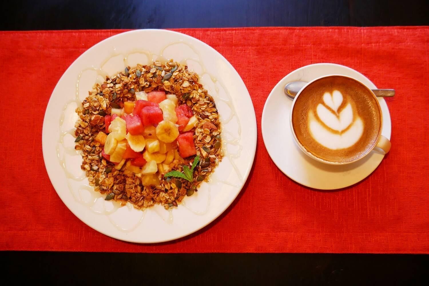 A plate of granola with fruits and a cup of coffee with latte art on a red tablecloth.