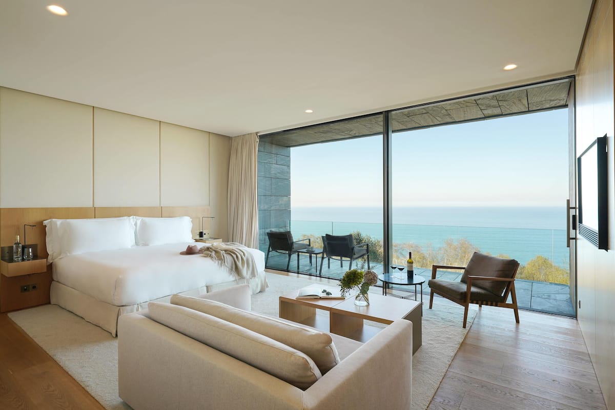 A modern bedroom with a large bed, a sofa, a coffee table, and a balcony with a view of the sea.