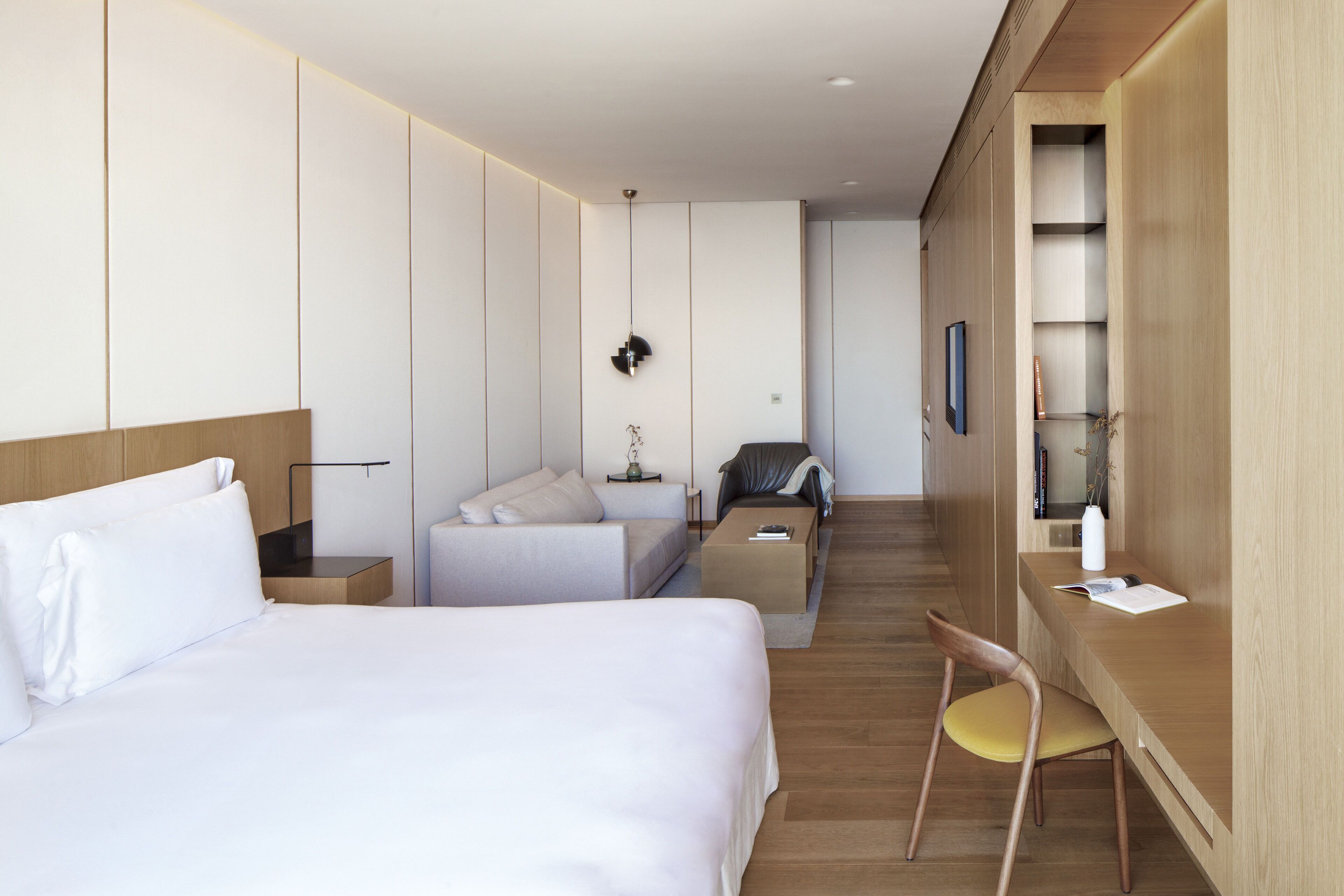 A modern hotel room with a large bed, a desk with a chair, a sofa, and a TV on the wall.
