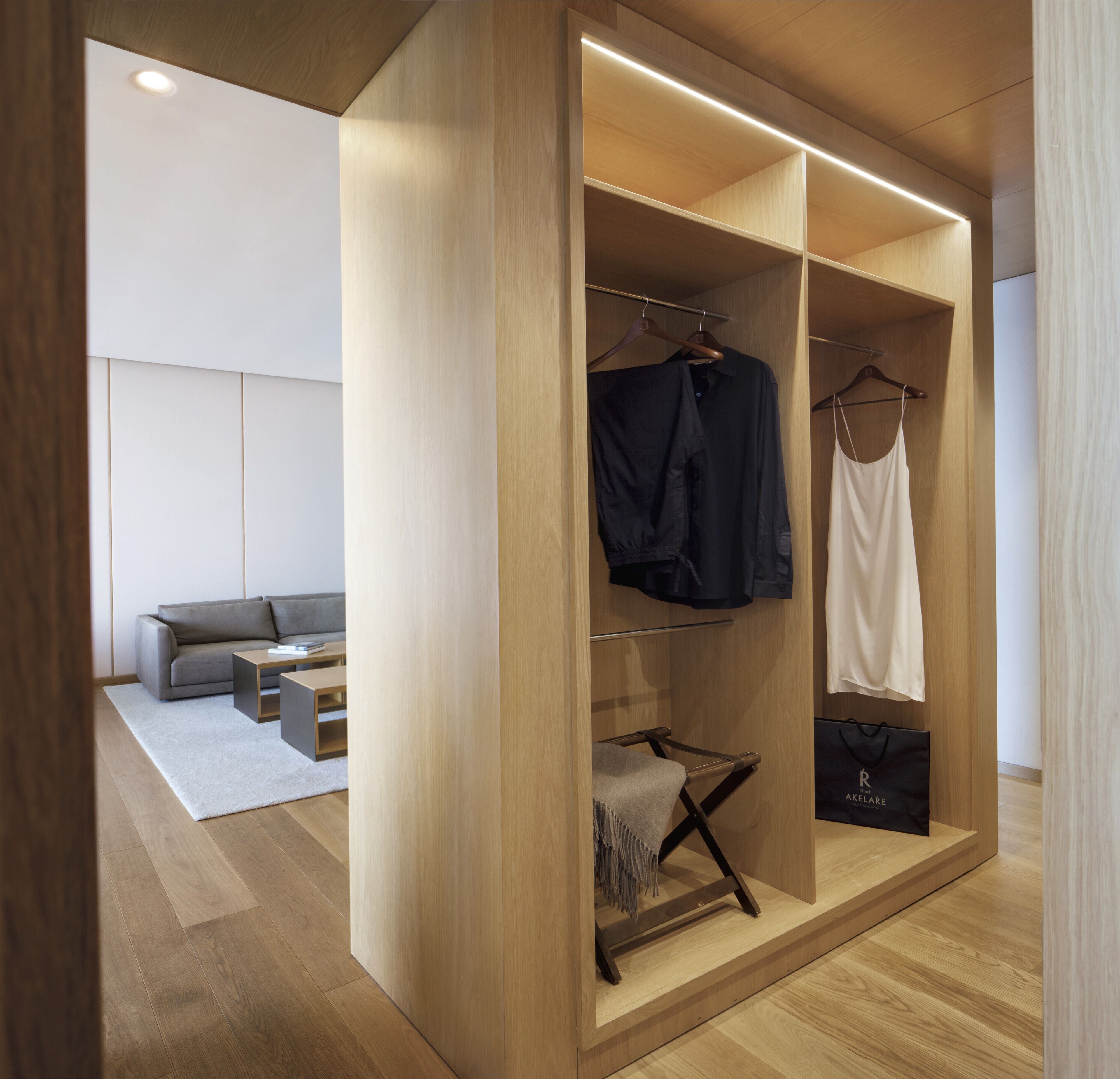 A modern, well-lit walk-in closet with wooden shelving and a hanging rod, featuring a grey sofa and a small table in the adjacent living area.