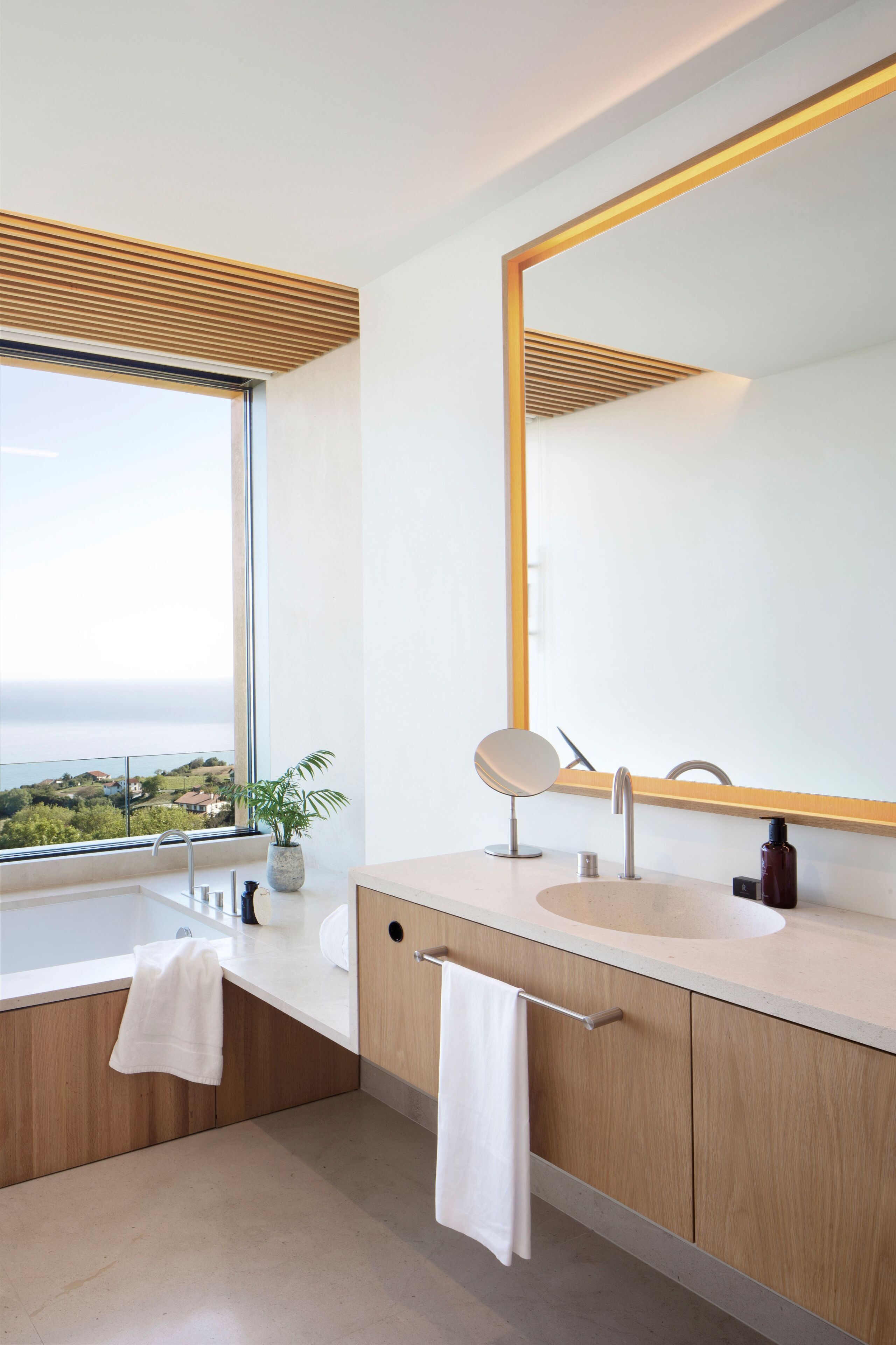 A modern bathroom with a large window, a bathtub, a sink, and a mirror.