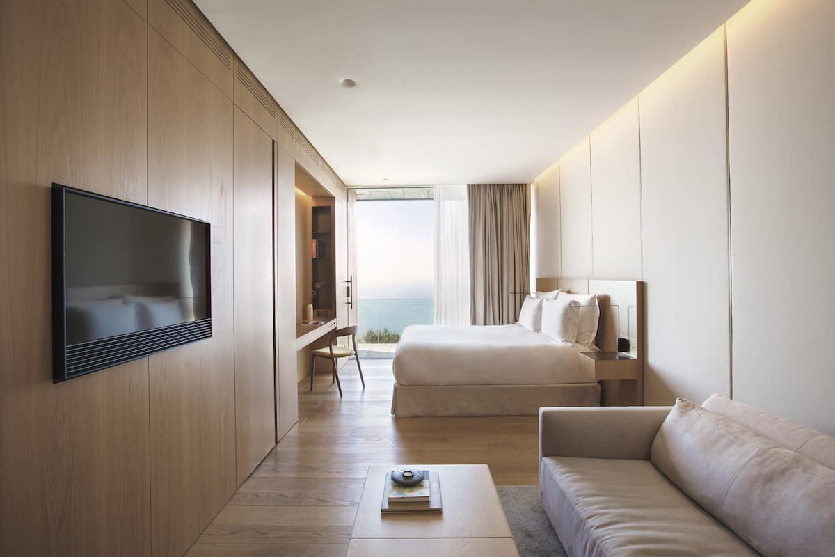 A modern hotel room with a large bed, a sofa, a TV mounted on the wall, and a view of the sea through the window.