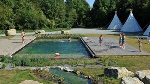 Een natuurzwembad met houten terrassen, omgeven door rotsen en begroeiing, met tipitentjes op de achtergrond.