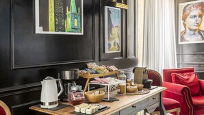 A room with a wooden table set with snacks, a coffee maker, and a kettle. There are framed artworks on the wall and a red chair in the corner. - Spagna Art & Suites - Domus Collection (Rome)