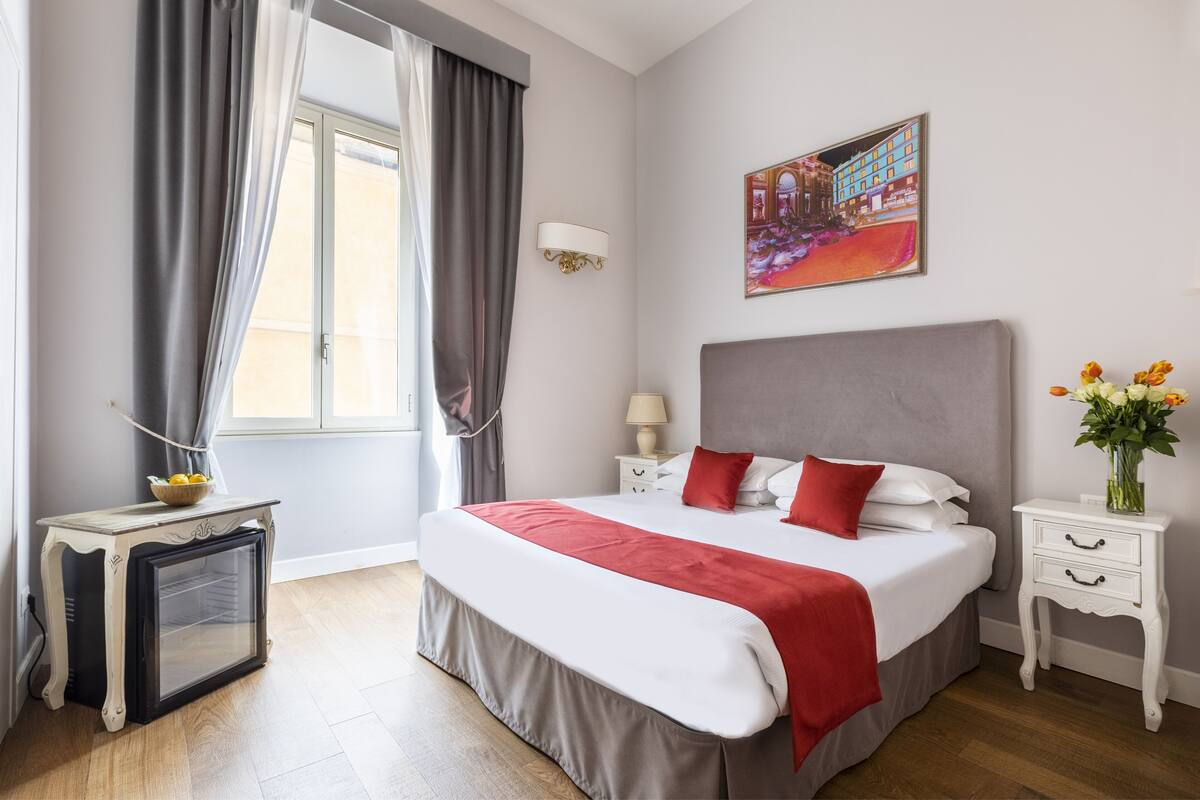 A hotel room with a bed, a bedside table, a nightstand, a window with curtains, a wall-mounted picture, and a small refrigerator.