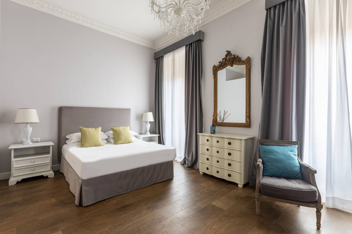 A bedroom with a large bed, a dresser, a chair, a mirror, and a chandelier.