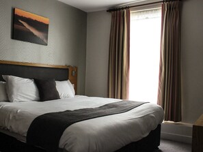 A hotel room with a large bed, a headboard, a painting on the wall, and a window with curtains. - Ye Olde Griffin (March)