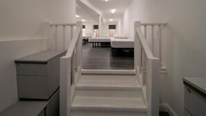 Un passadís estret amb parets blanques i una escala de fusta que condueix a una habitació amb un llit i una finestra.
